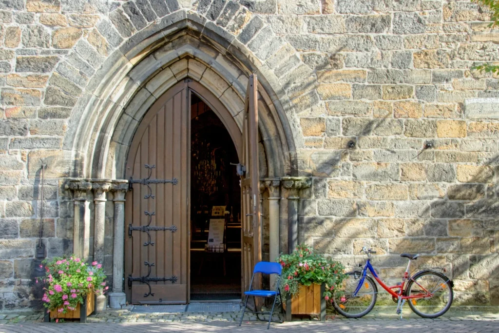 Geöffnete Kirchentür mit Blumenkästen, Klappstuhl und an die Kirchenmauer angelehntem Fahrrad